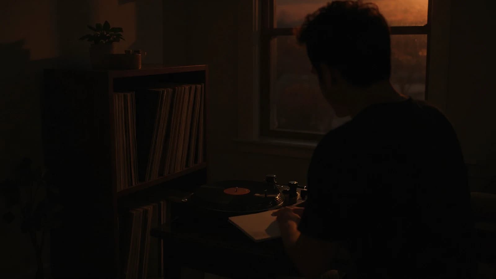 A collector writing notes beside a turntable and a shelf of records in a softly lit room.
